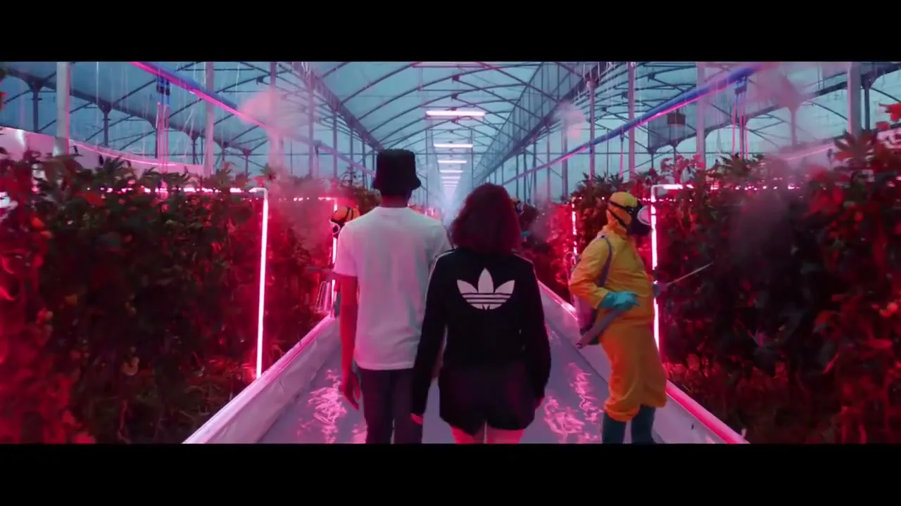 A young man and woman walk away from the camera down a long, narrow path in a futuristic greenhouse with red-lit plants and people in hazmat suits spraying mist.