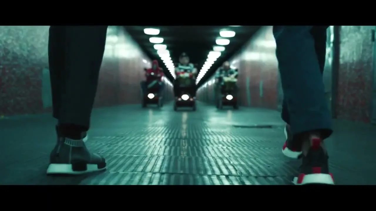 Two people walk away from the camera in a dark, tiled tunnel, while three others on mobility scooters approach them in the distance.