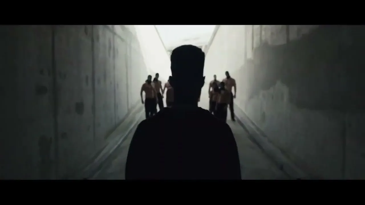 A young man, silhouetted, walks away from the camera down a dark concrete tunnel, as a group of men approach him from the distance.