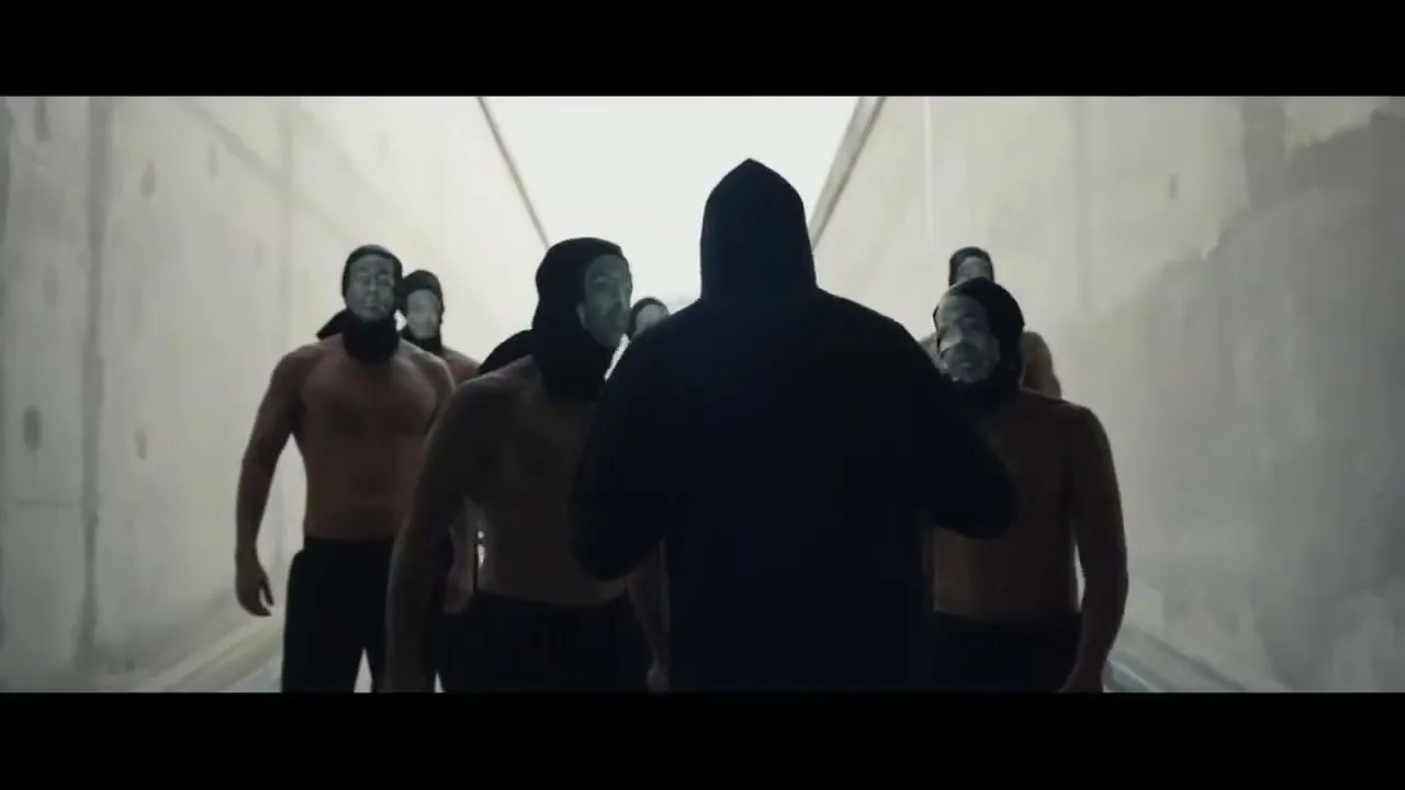 A hooded figure in silhouette faces a group of shirtless men wearing masks, standing in a stark, concrete tunnel.
