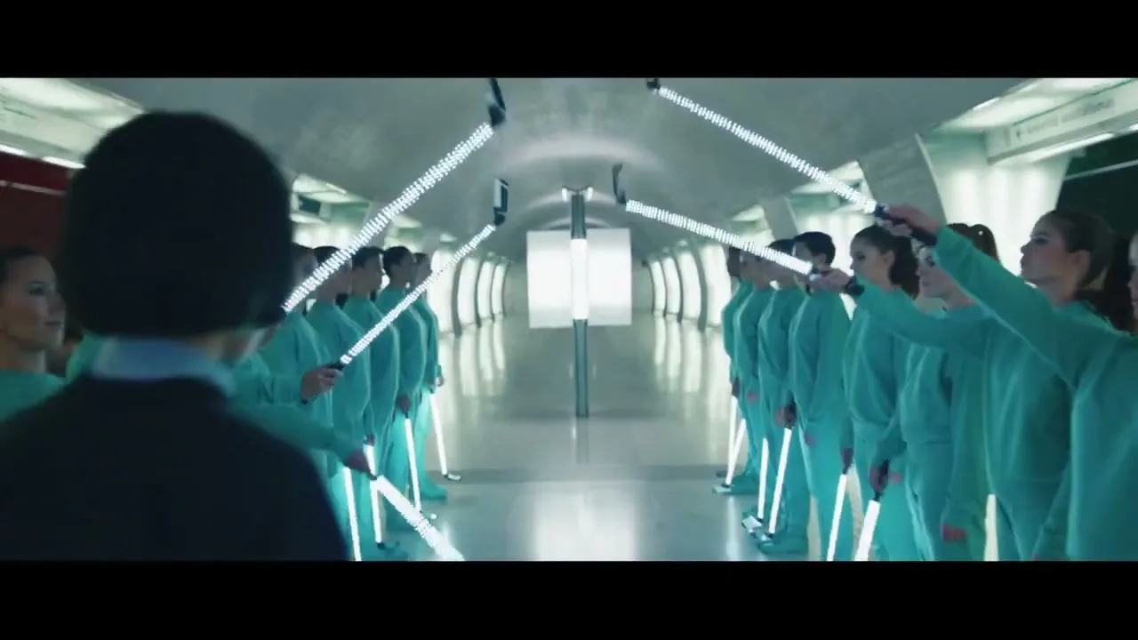 A young woman with short dark hair stands with her back to the camera, facing two lines of young women in matching teal tracksuits holding glowing light sticks in a futuristic tunnel.