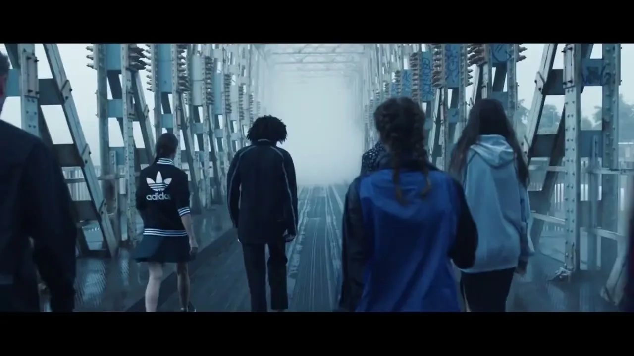A diverse group of young people walk away from the camera across a misty, industrial bridge, disappearing into a bright white fog.