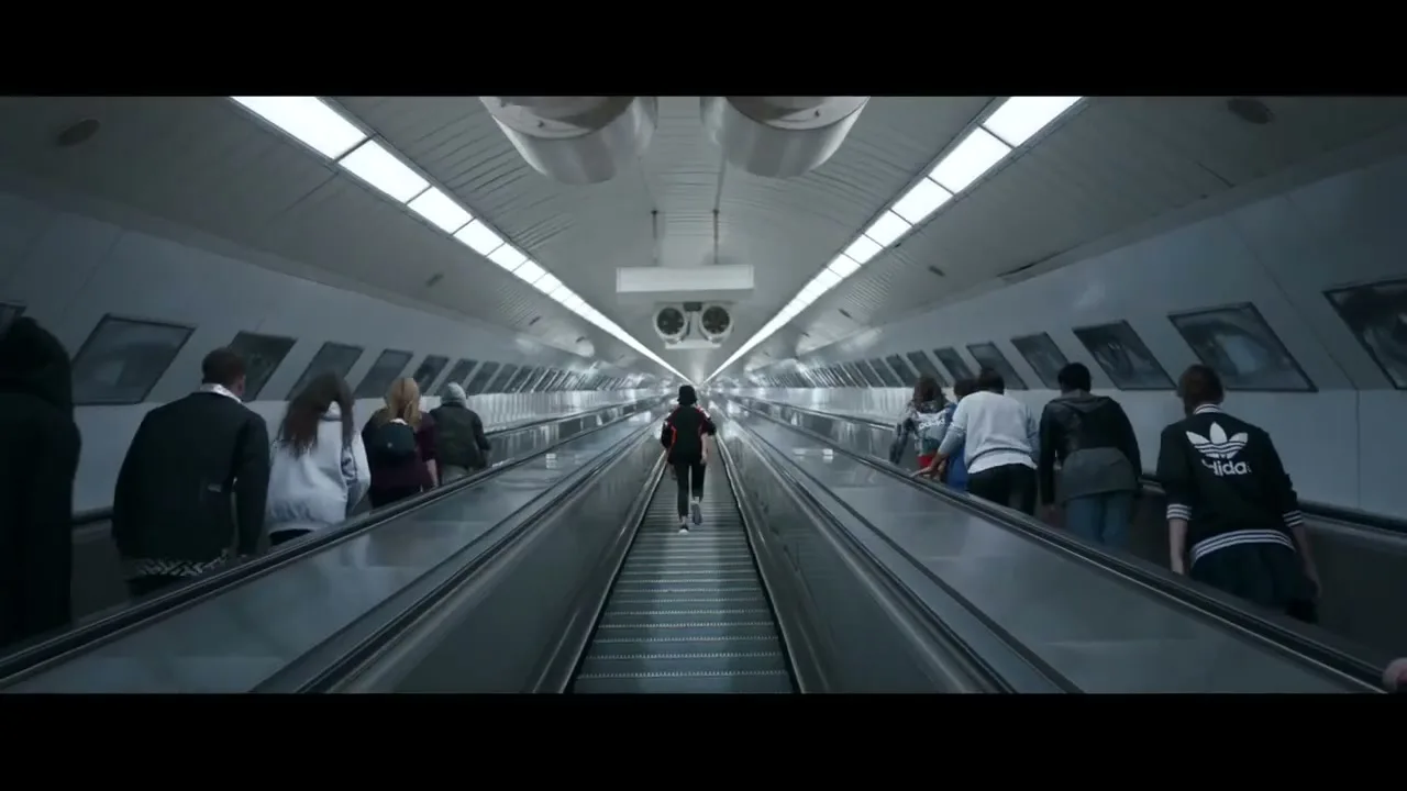 A young person in a black and red tracksuit runs up a long, brightly lit escalator in a subway station, while other people stand on adjacent escalators.