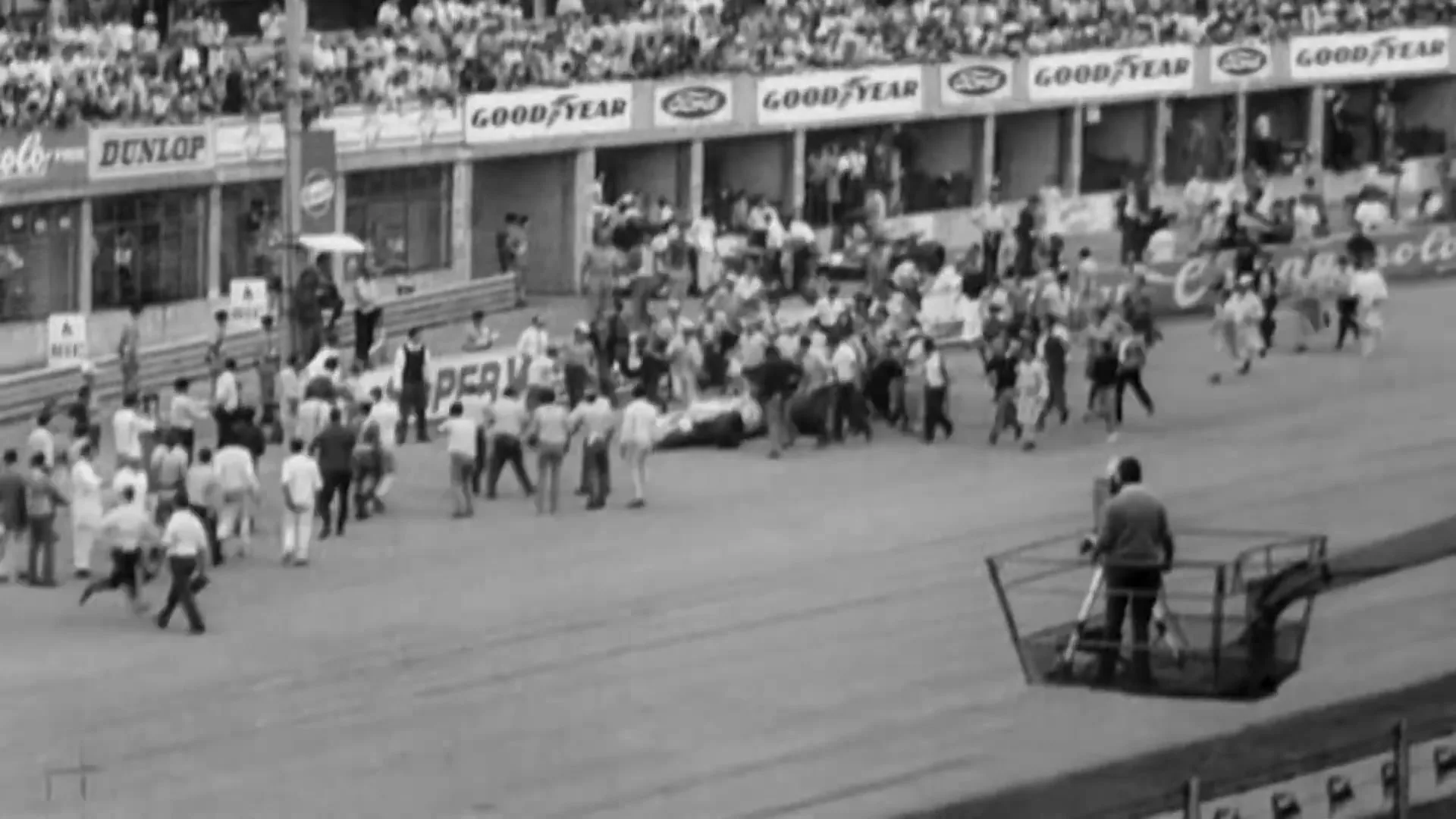 A black and white archival shot shows a crowd of people running onto a race track after a car crash, with a cameraman filming from a raised platform.