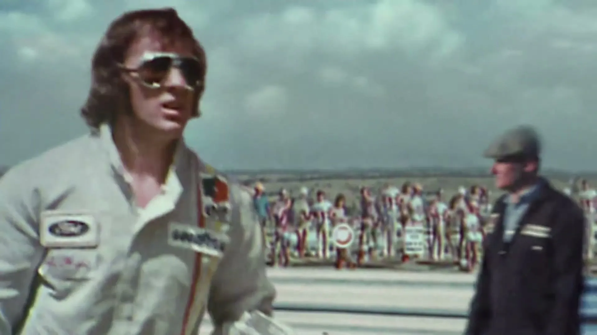 A young man in a racing suit and sunglasses walks past a crowd of people at a racetrack, then turns and walks towards a building.
