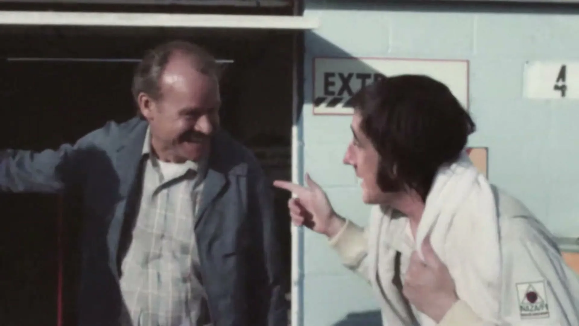 A male mechanic in a blue jacket and a male race car driver in a white suit are laughing and talking outside a garage.