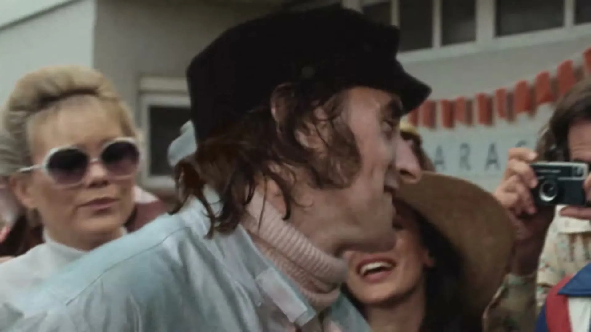 A male race car driver in a black cap and light blue jacket smiles and laughs with a female fan in a straw hat, surrounded by a small crowd outdoors.