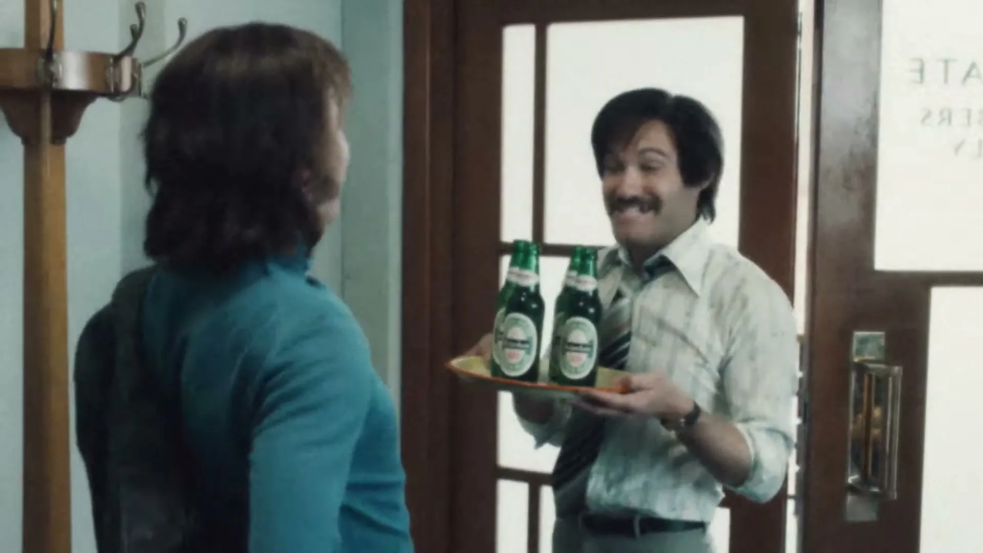 A man with a mustache, wearing a striped shirt and tie, holds a tray of Heineken bottles for a woman in a blue top, in an interior setting.