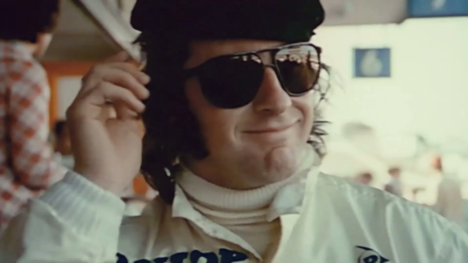 A man in a white racing suit, black cap, and sunglasses smiles, then turns his head to the right, at a racetrack.