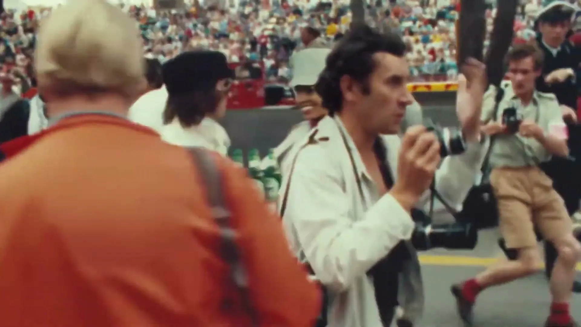 A male photographer in a white jacket and black shirt holds a camera, looking to the right, surrounded by a crowd at a race track.