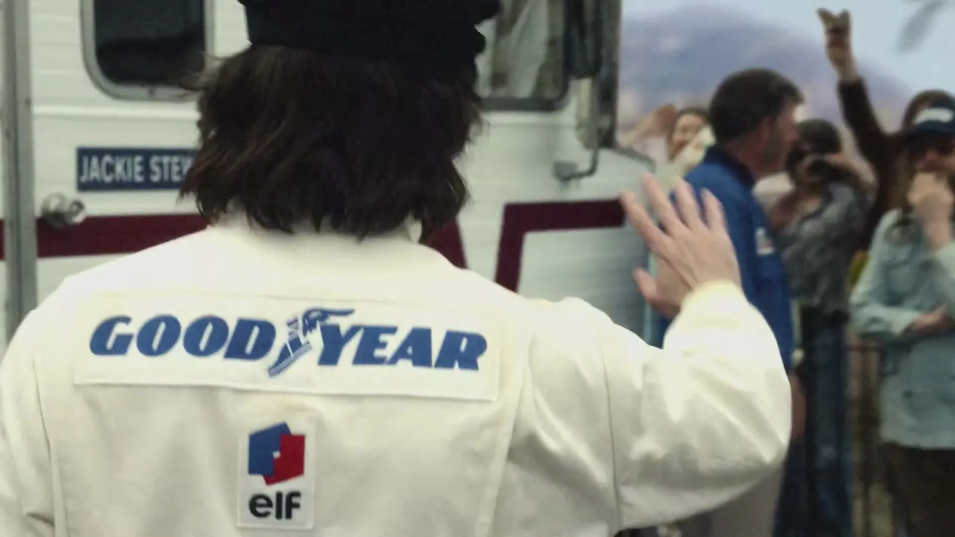 A man in a white racing suit with "GOOD YEAR" on the back waves to a small crowd of people standing next to a white RV.