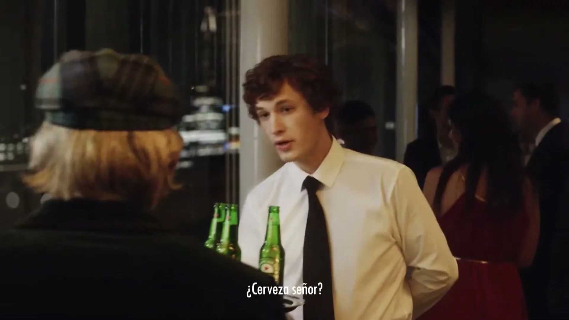 A young male waiter in a white shirt and black tie offers a tray of Heineken bottles to an older man at a formal evening event.