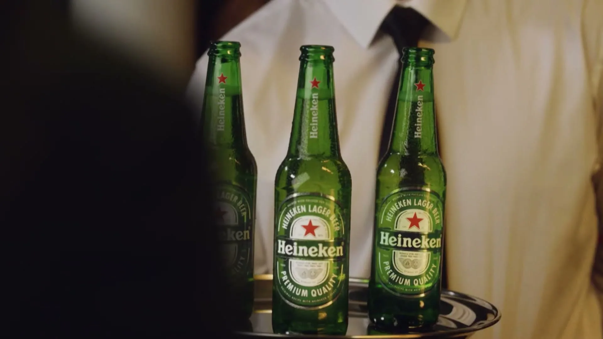Three green Heineken beer bottles with condensation are presented on a silver tray by a waiter in a white shirt.
