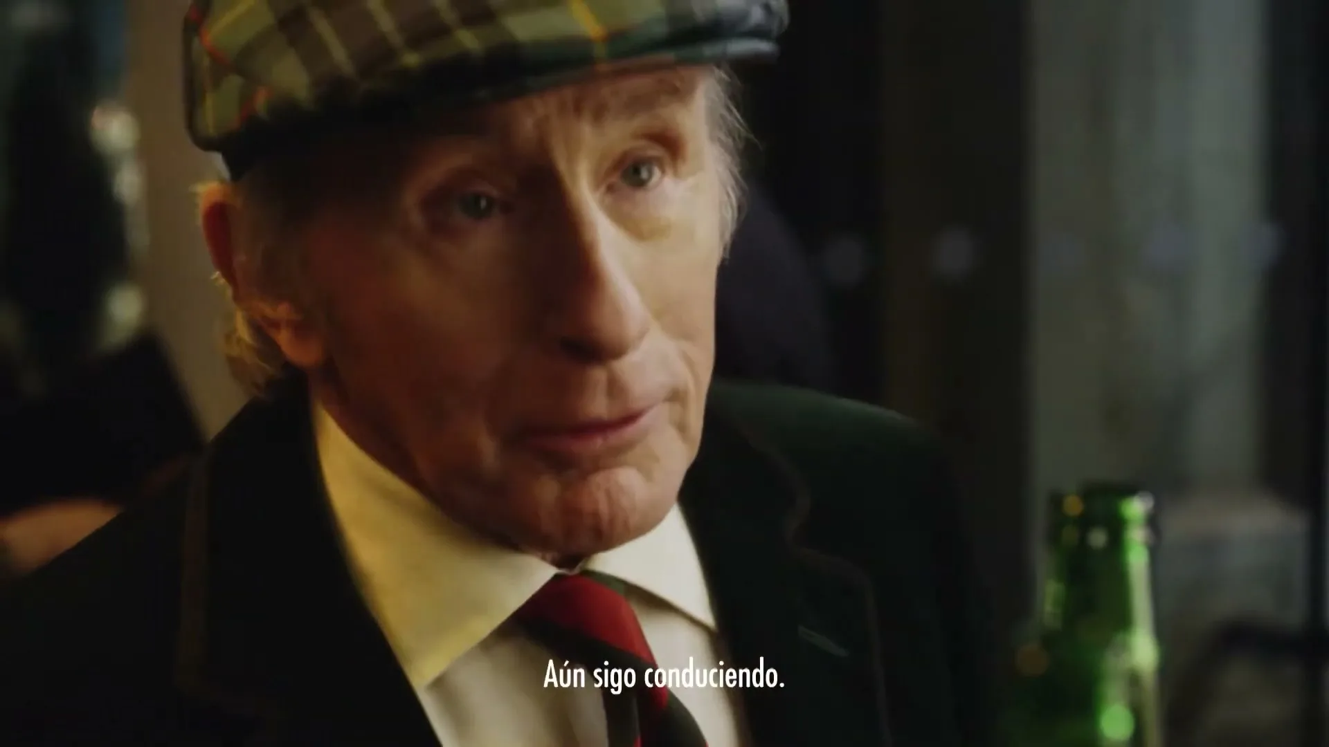 An older man in a plaid cap and suit speaks, then smiles and adjusts his cap, with a green bottle visible beside him in a dimly lit interior.