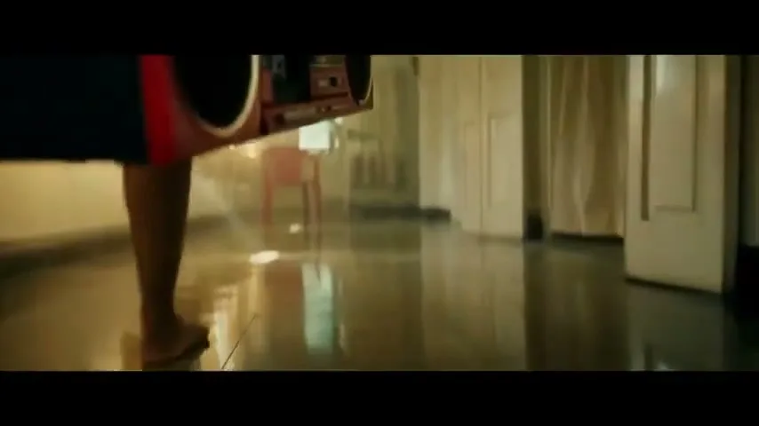 A person walks barefoot across a shiny floor, carrying a red boombox, in a dimly lit, empty room.