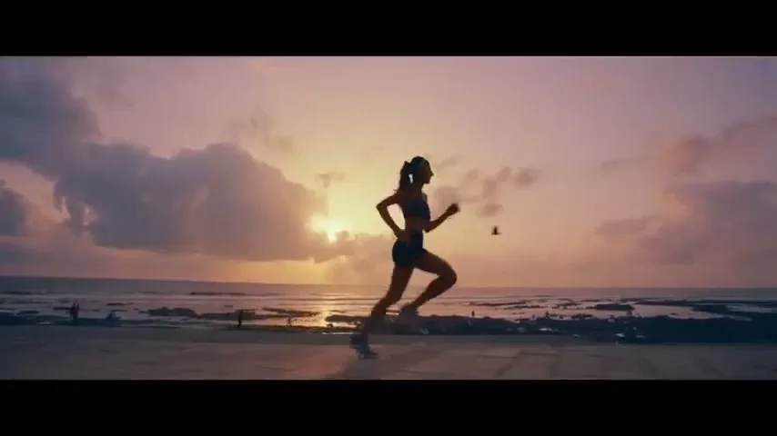 A female athlete runs in silhouette along a beach at sunset, with the ocean and a cloudy sky in the background.