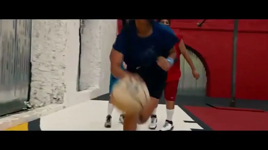 A female basketball player in a blue shirt dribbles the ball while another player in red defends her on an indoor court.