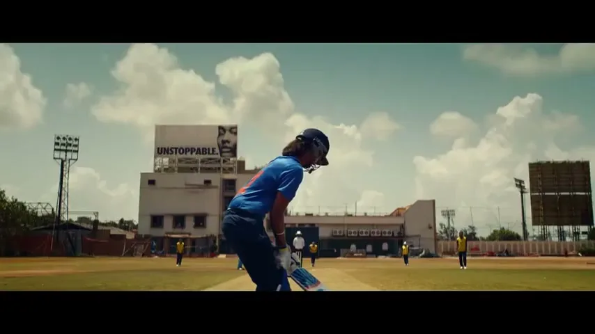 A female cricket player in blue uniform and helmet stands ready to bat on a sunny day at a cricket ground, with a large billboard in the background.
