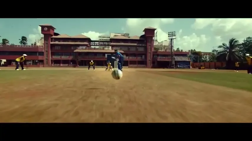 A wide shot shows a cricket player in blue uniform hitting a ball on a dusty field, with other players and a red building in the background.