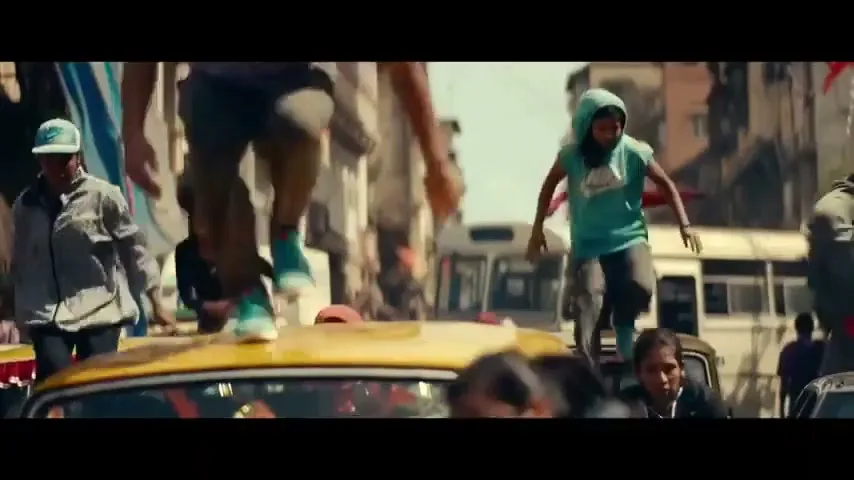 A group of young people, including a male in a purple shirt and a female in a teal hoodie, are running and jumping over yellow taxis in a busy street.