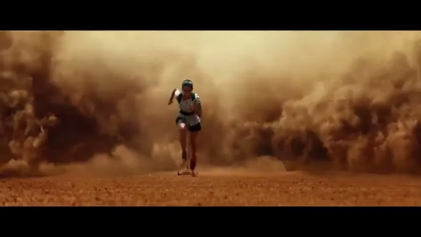 A female athlete runs towards the camera through a dusty, arid landscape, with large clouds of dust rising behind her.