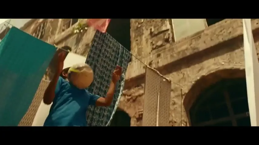 A young boy in a blue shirt plays with a soccer ball in an alleyway with clotheslines and old buildings.