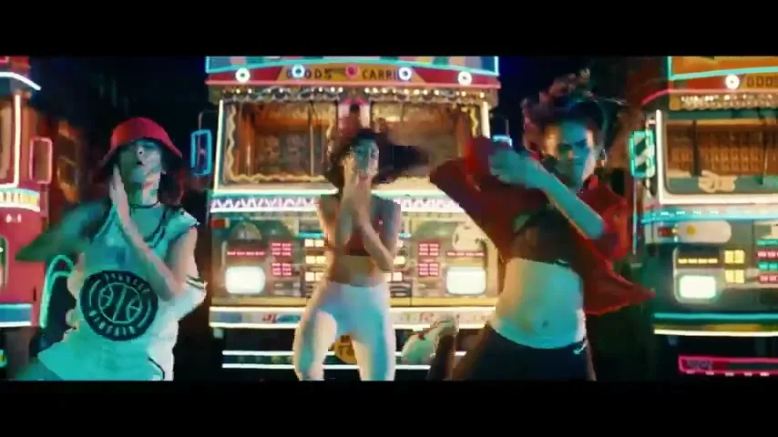 Four young women in athletic wear dance energetically in front of brightly lit trucks at night.