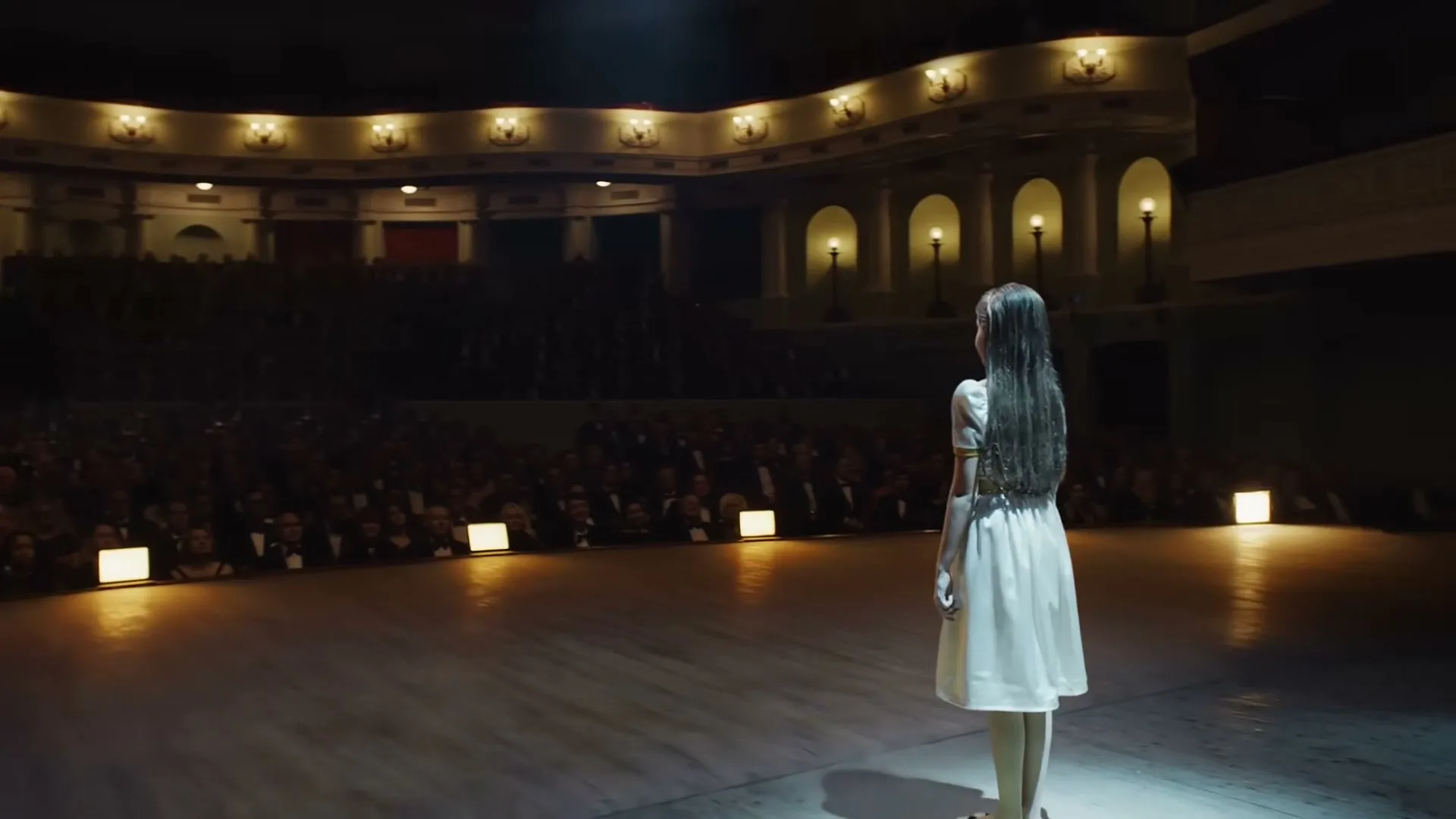 A young girl in a white dress with a gold headband walks from a dark backstage area onto a brightly lit stage, facing a large, dark audience.