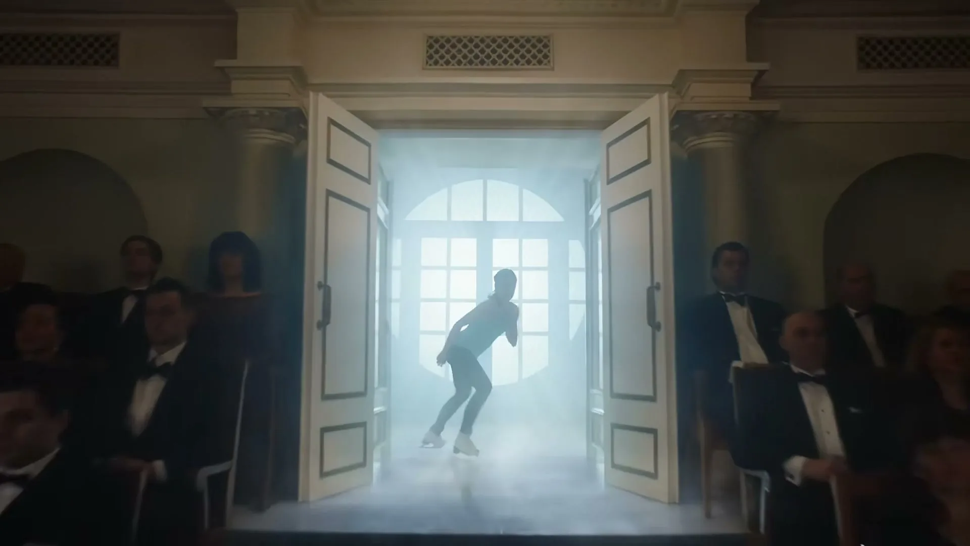 A young female athlete in a blue tank top and black leggings runs out of a brightly lit doorway onto a dark stage, where an audience in formal wear watches her.