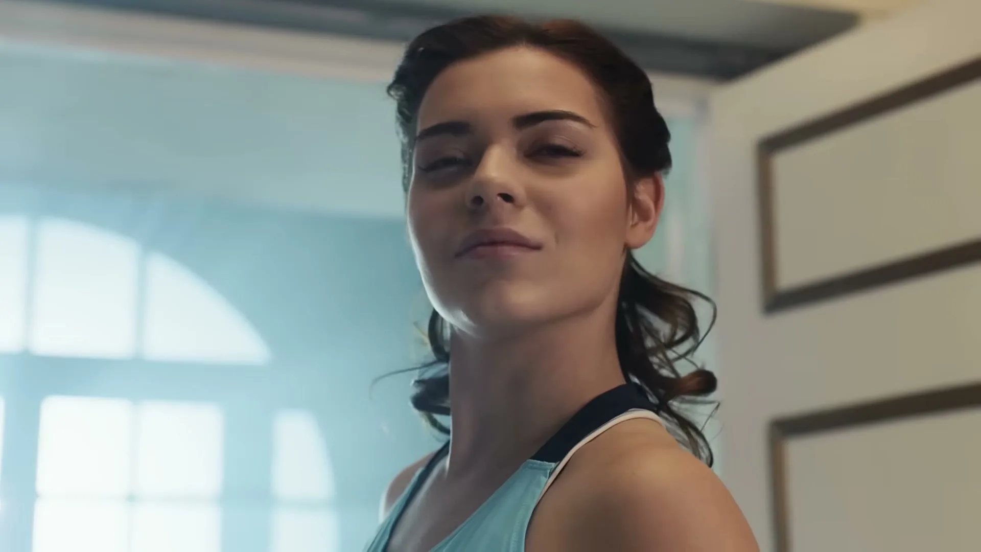 A young woman with dark hair in a light blue sports bra looks directly at the camera with a determined expression in a bright, airy room.