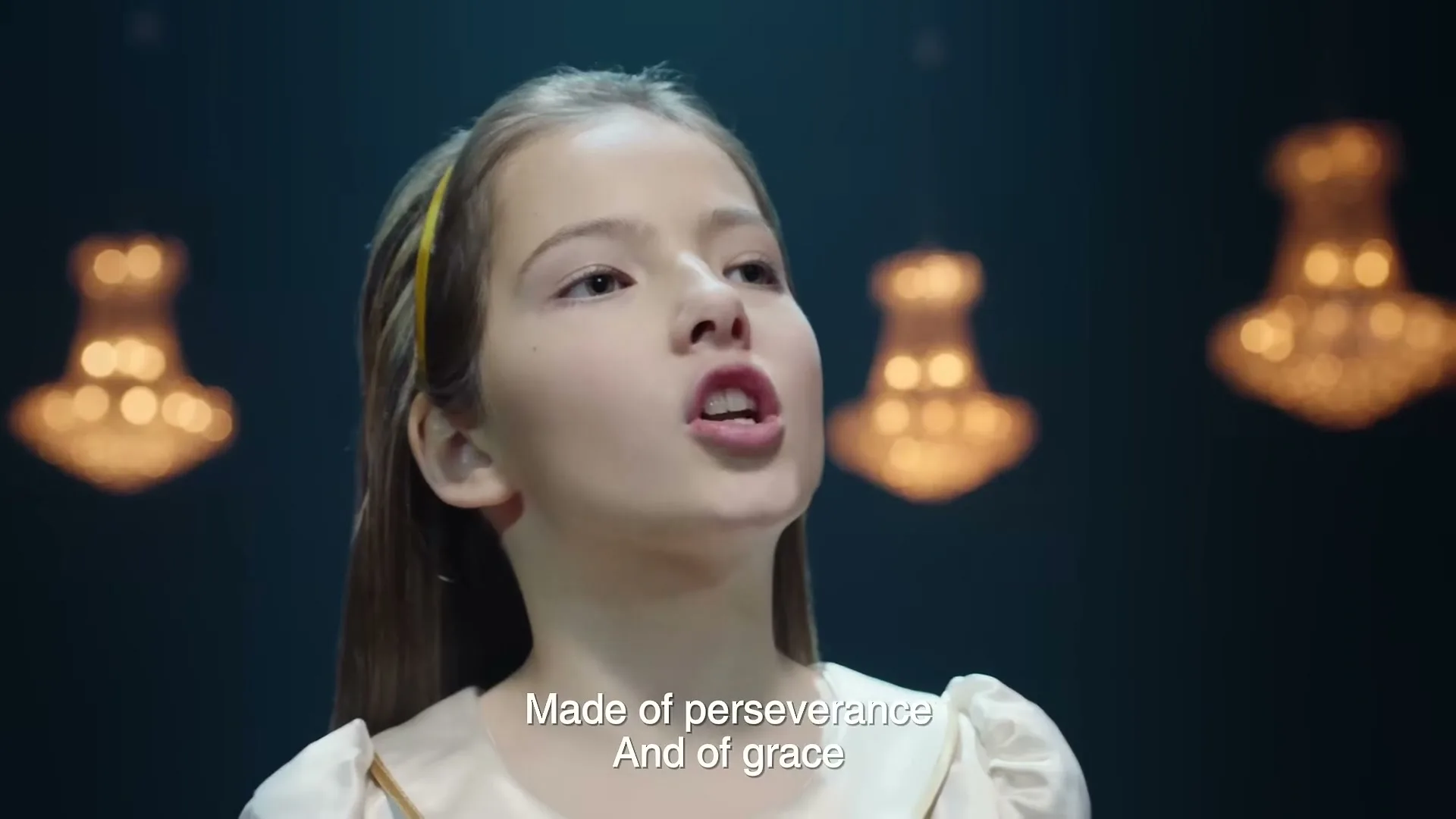 A young girl with light brown hair and a yellow headband sings passionately into the camera, with blurred chandeliers in the background.