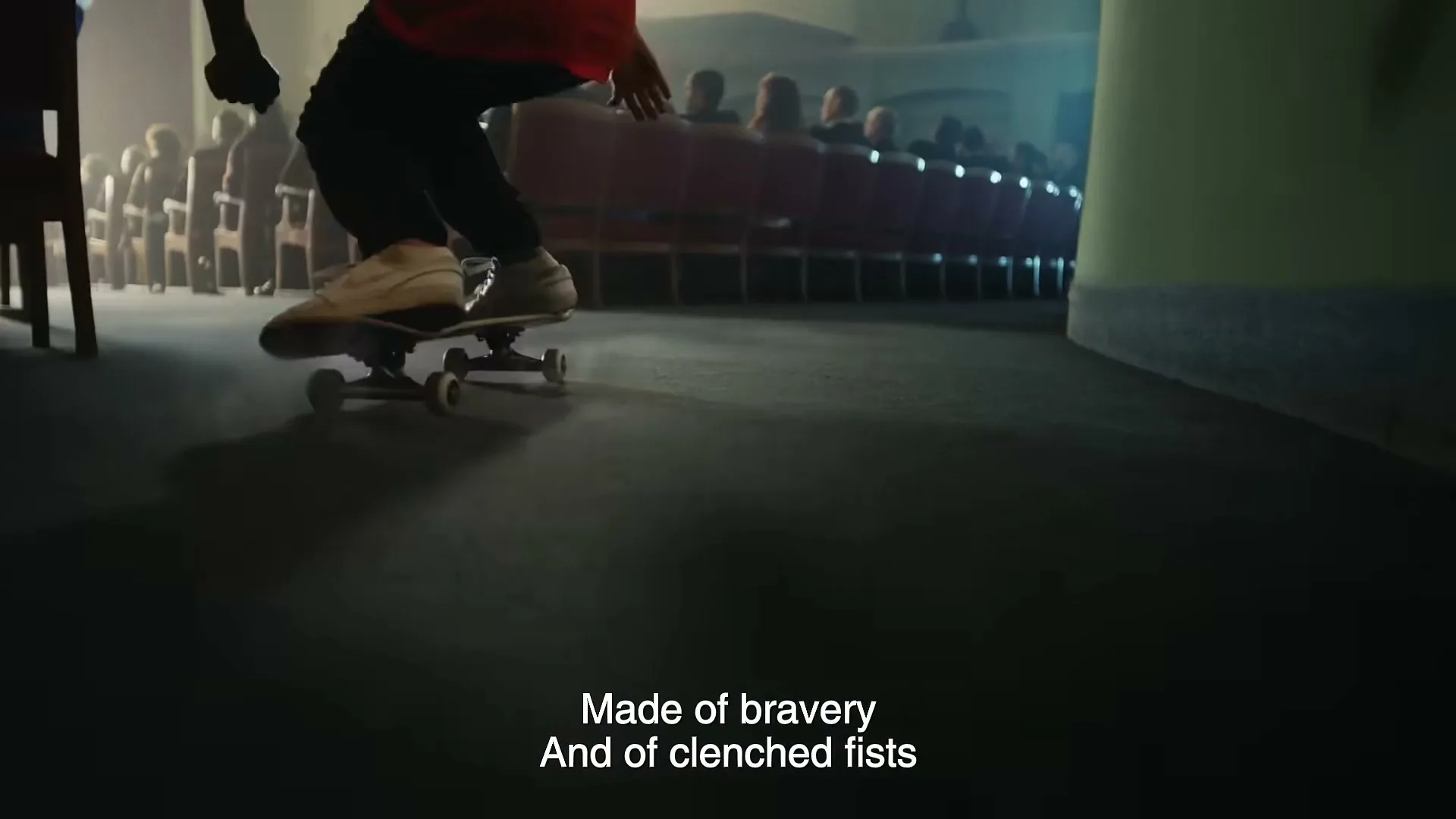 A young person on a skateboard glides across the floor of a dimly lit auditorium, with an audience seated in the background.