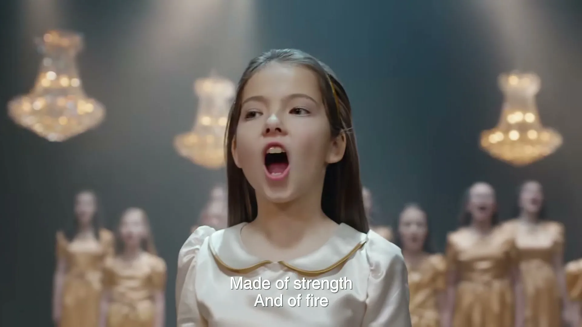 A young girl with long brown hair and a white dress sings passionately, surrounded by other girls in gold dresses, under chandeliers.