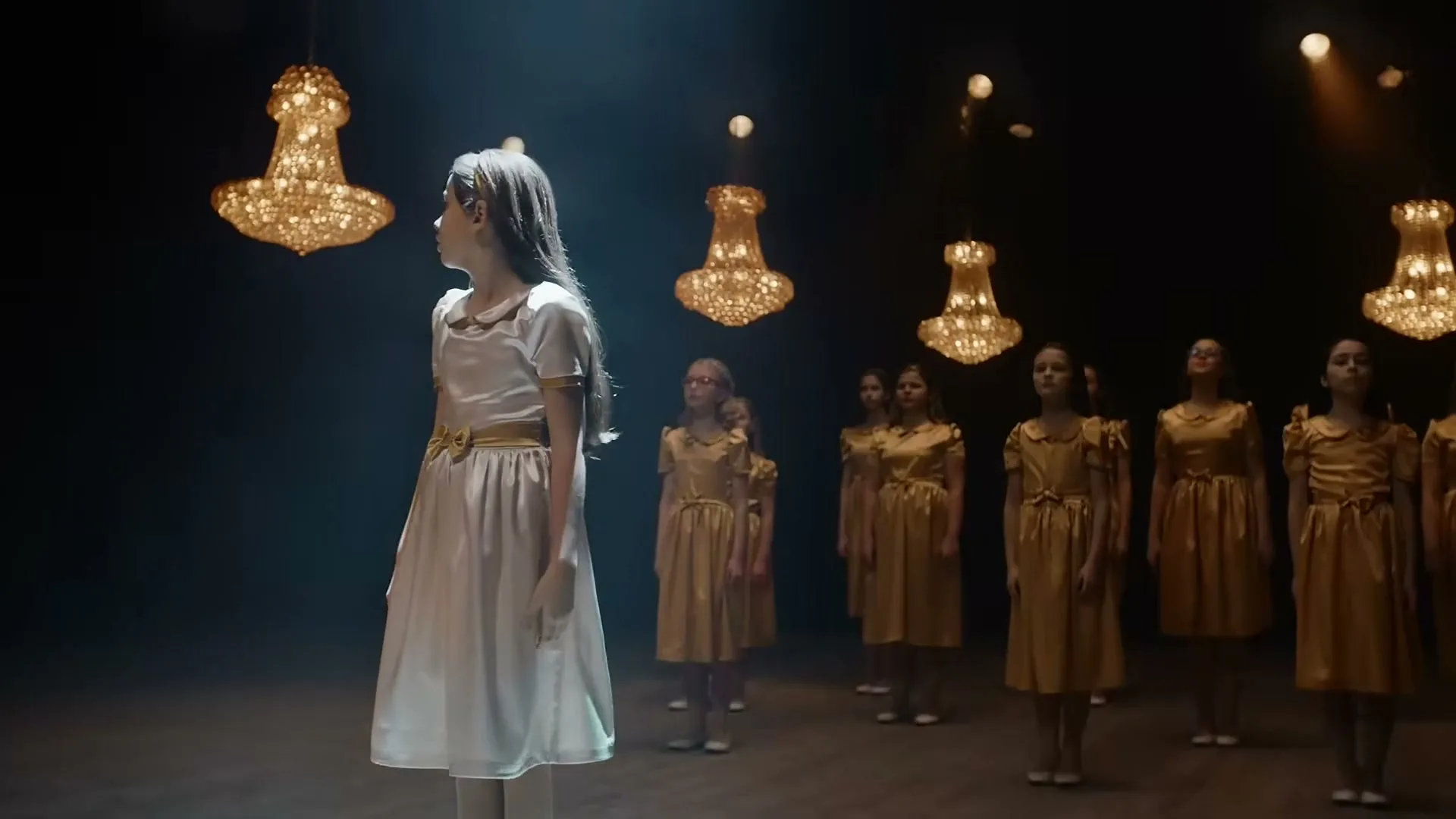 A young girl in a white dress stands center stage, looking up, surrounded by other girls in gold dresses under chandeliers in a dark theater.