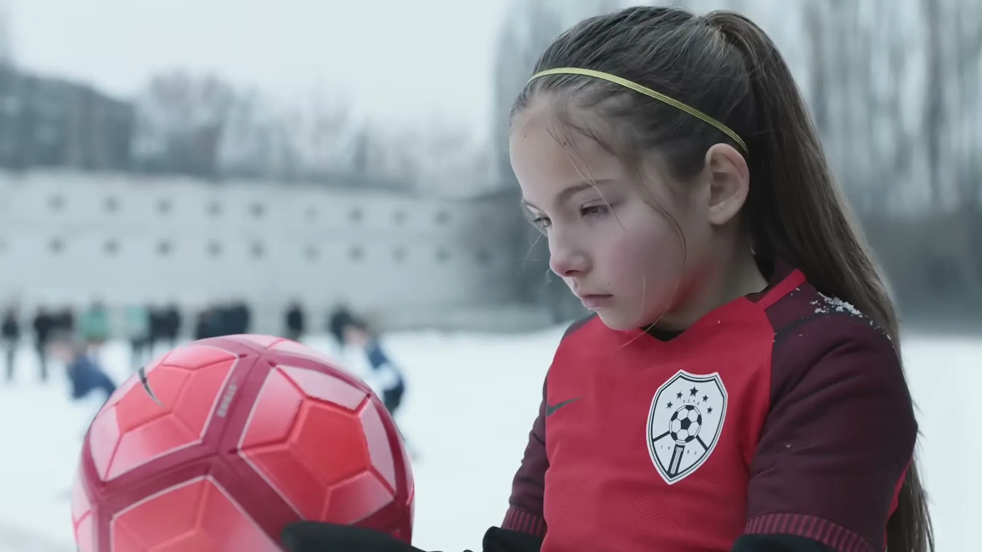 A young girl in a red soccer jersey holds a red soccer ball, looking determined on a snowy field with other players in the background.