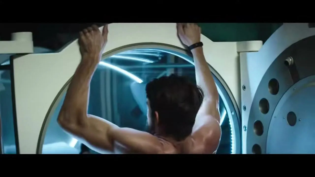 A muscular man with dark hair, seen from behind, hangs from the top of a hyperbaric chamber, his back muscles flexed.