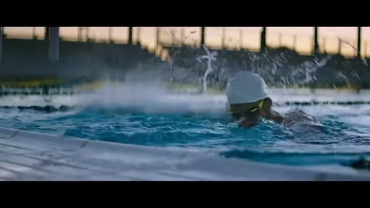 A male swimmer with a beard, wearing a white swim cap and goggles, swims butterfly stroke in an outdoor pool at dusk.