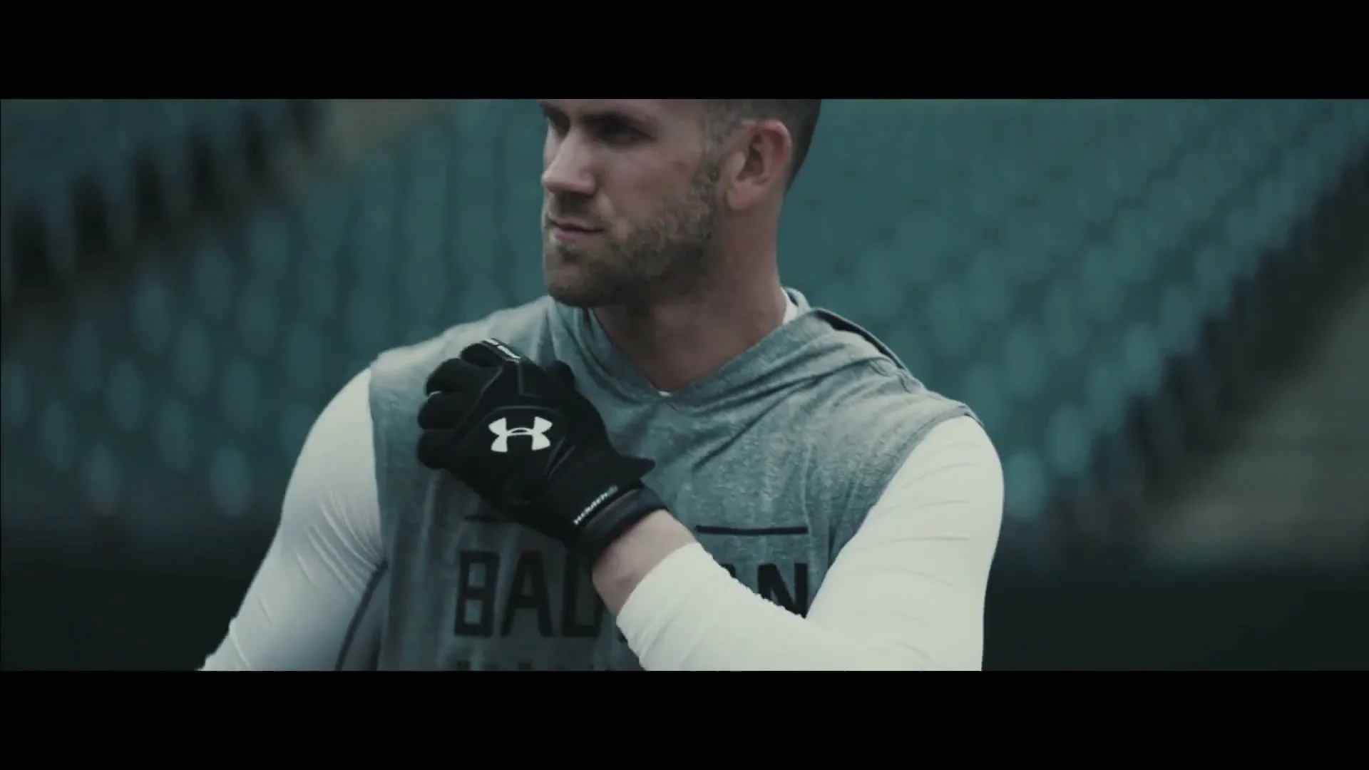 A male athlete in his 20s, wearing a grey hoodie and white long-sleeve shirt, stretches his arm in a stadium.