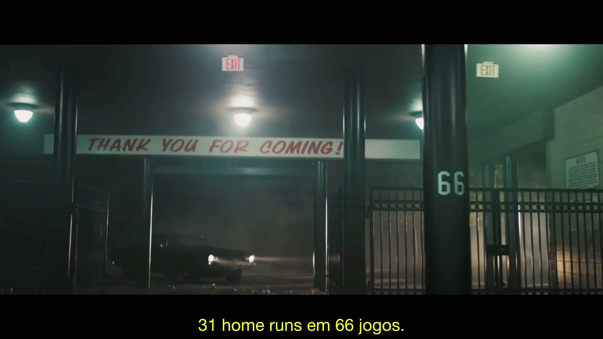 A dark car with headlights on drives slowly through a stadium tunnel at night, passing a "Thank You For Coming!" sign and a fence with the number 66.