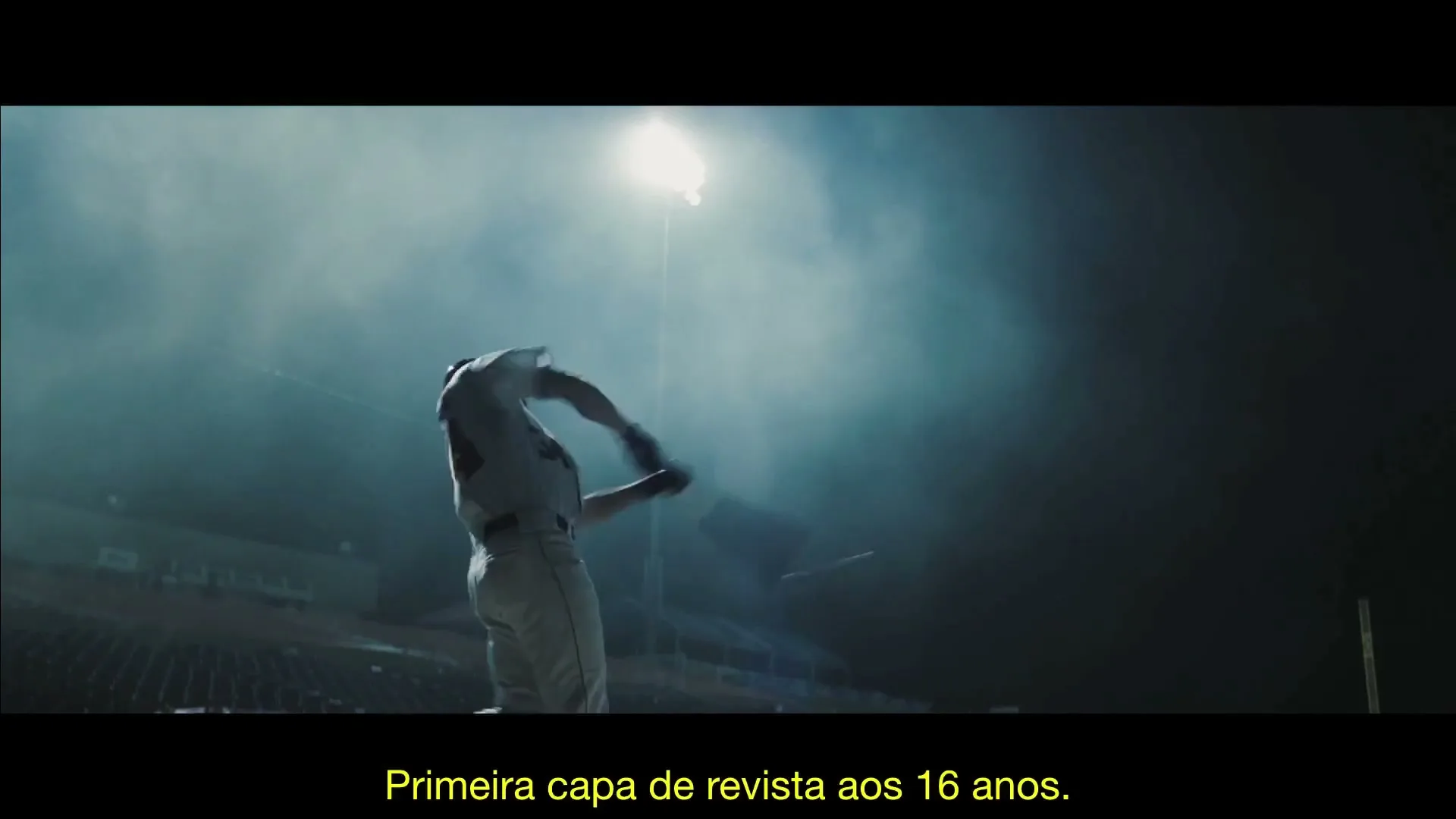 A male baseball player in a white uniform swings a bat under stadium lights at night.