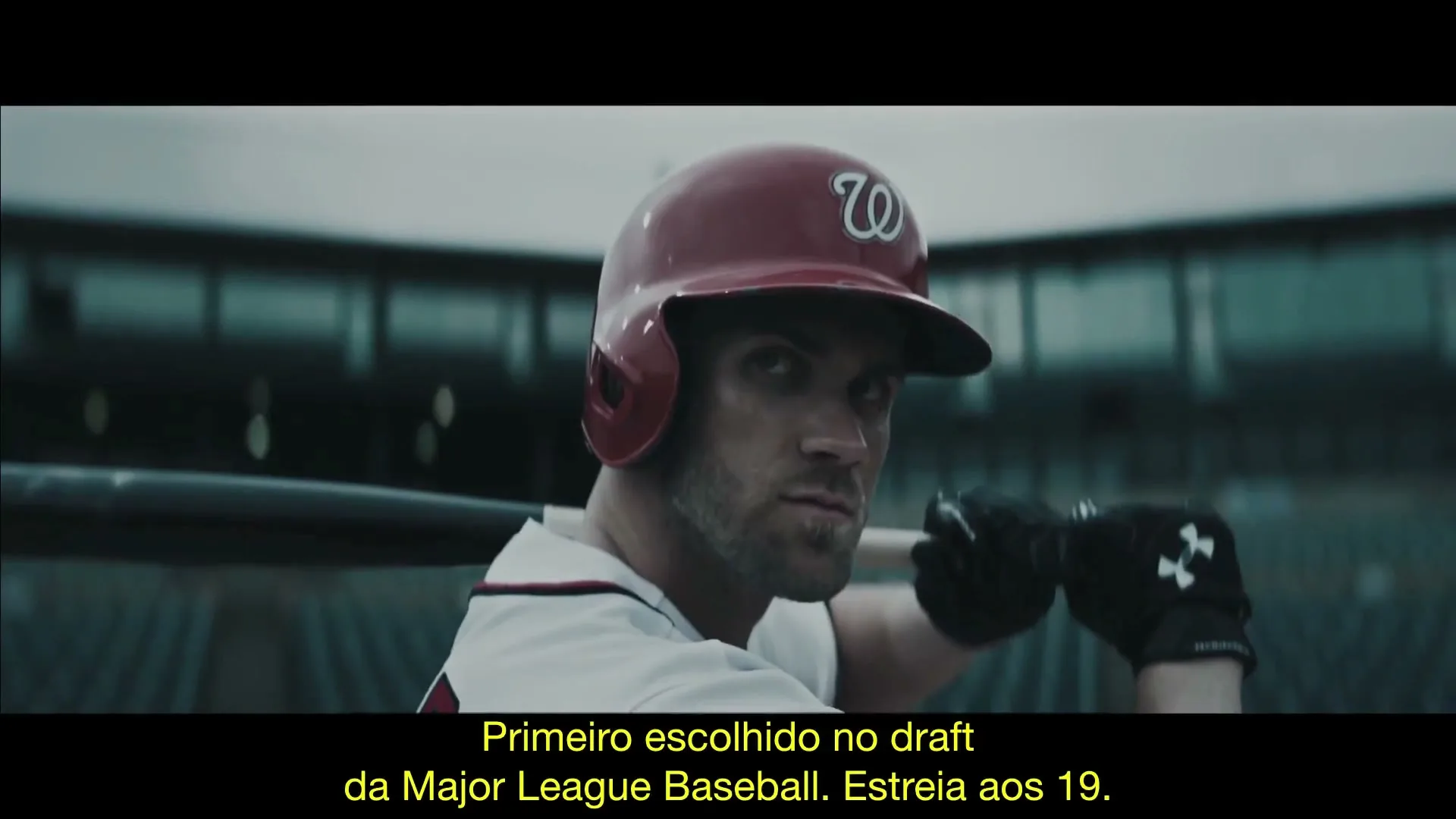 A male baseball player in a red helmet and white jersey holds a bat, looking intensely forward in a stadium.