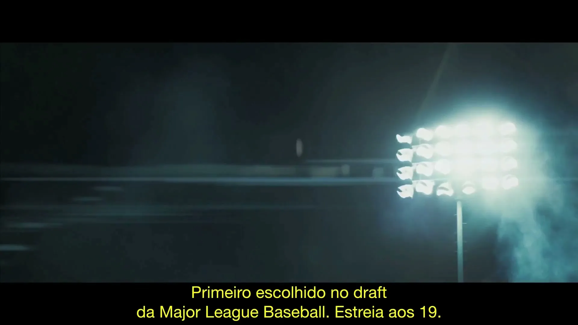 A wide shot of a baseball stadium at night, with a single player running on the field and bright stadium lights illuminating the scene.