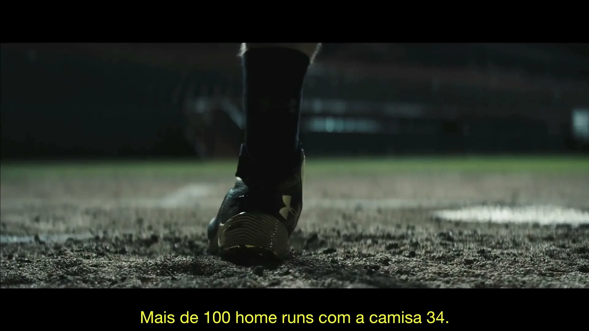 A baseball player's cleated foot, wearing a dark sock and gold-accented shoe, steps onto a dirt baseball field at night.