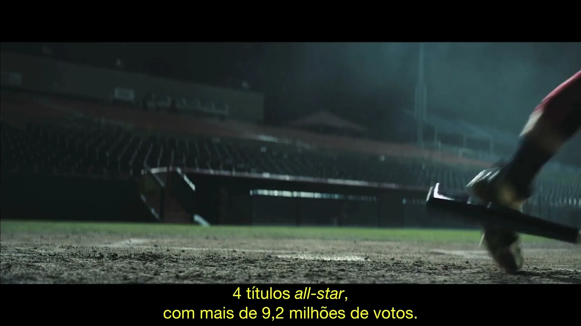 A baseball player in dark uniform and gold cleats runs across a dirt field, dropping a bat, with empty stadium seats in the background at night.