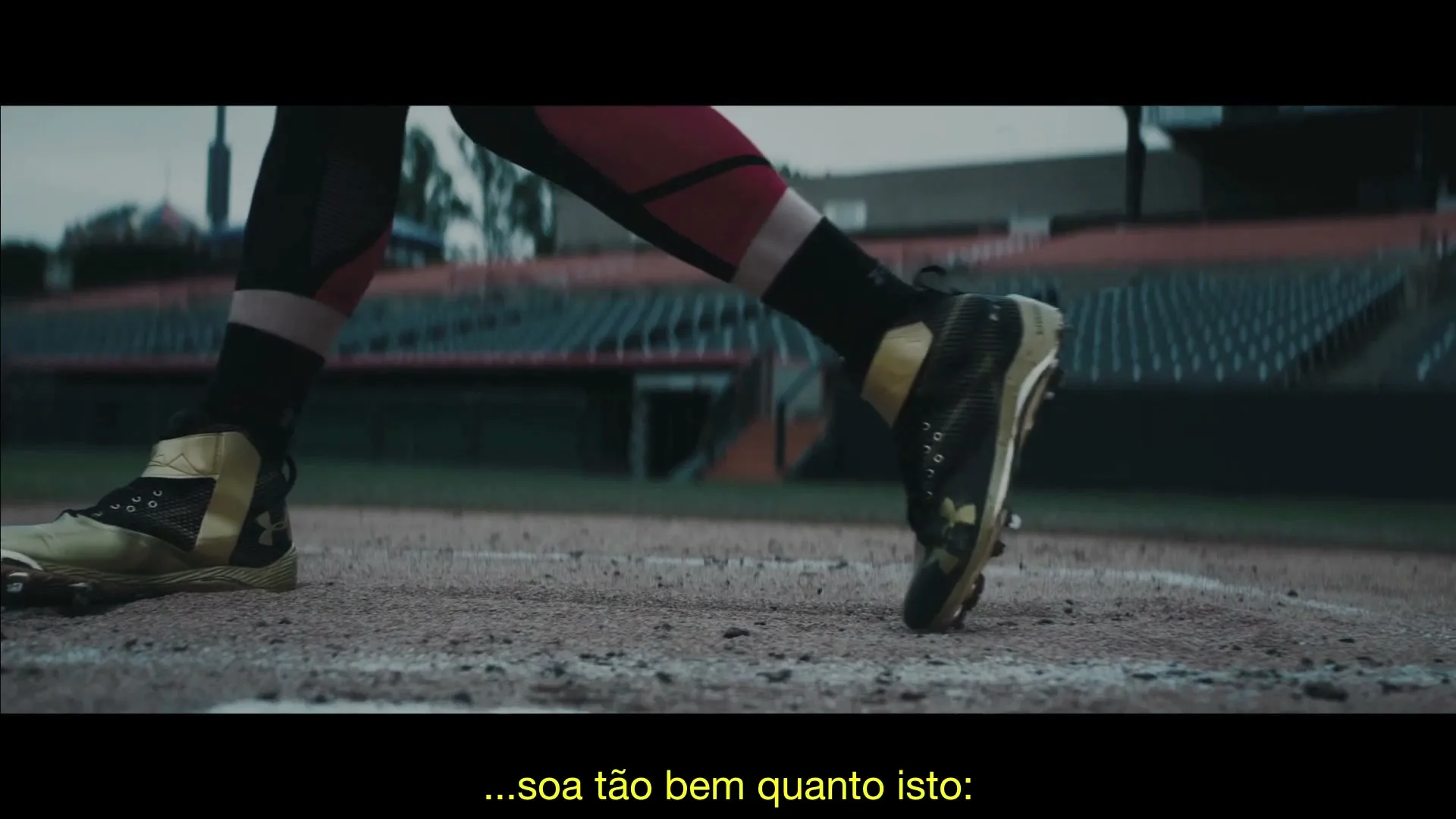 A baseball player's feet in gold and black cleats dig into the dirt of a baseball field, with empty stadium seats in the background.