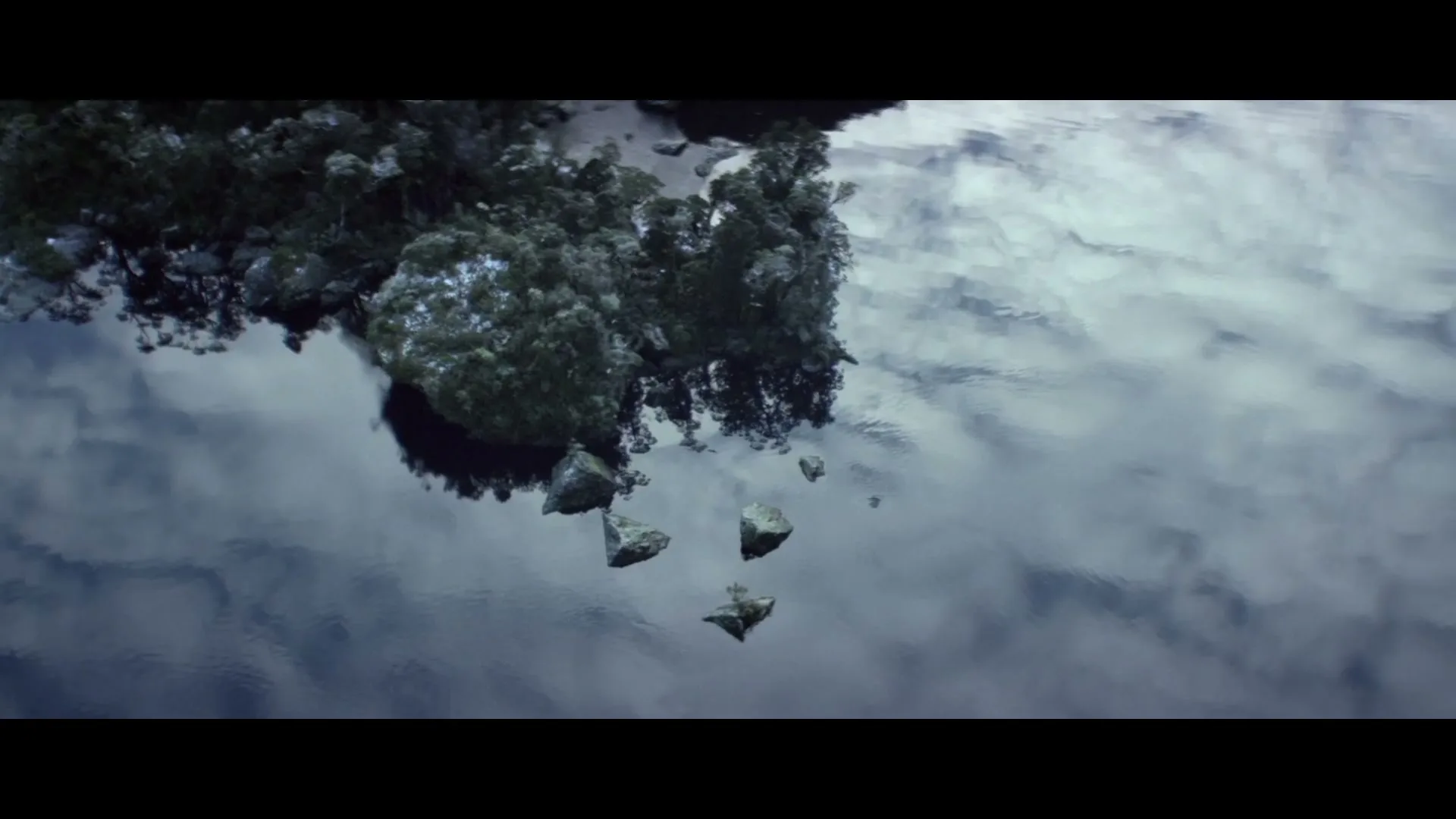 An aerial shot shows a small, rocky island covered in green trees, reflected in the still water of a lake under a cloudy sky.