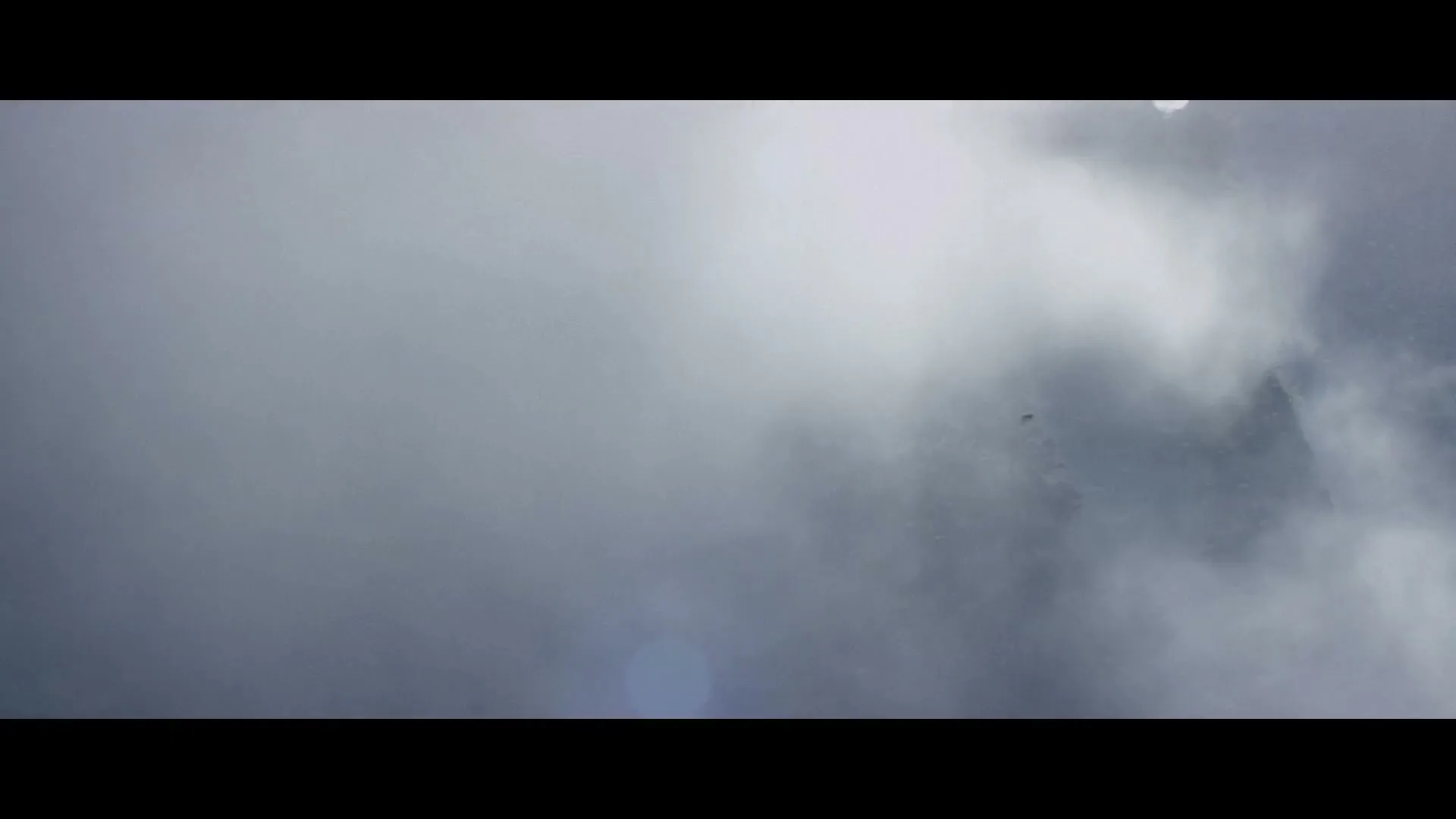 A wide shot of a dense, misty cloudscape with a faint outline of a mountain and trees emerging from the fog.