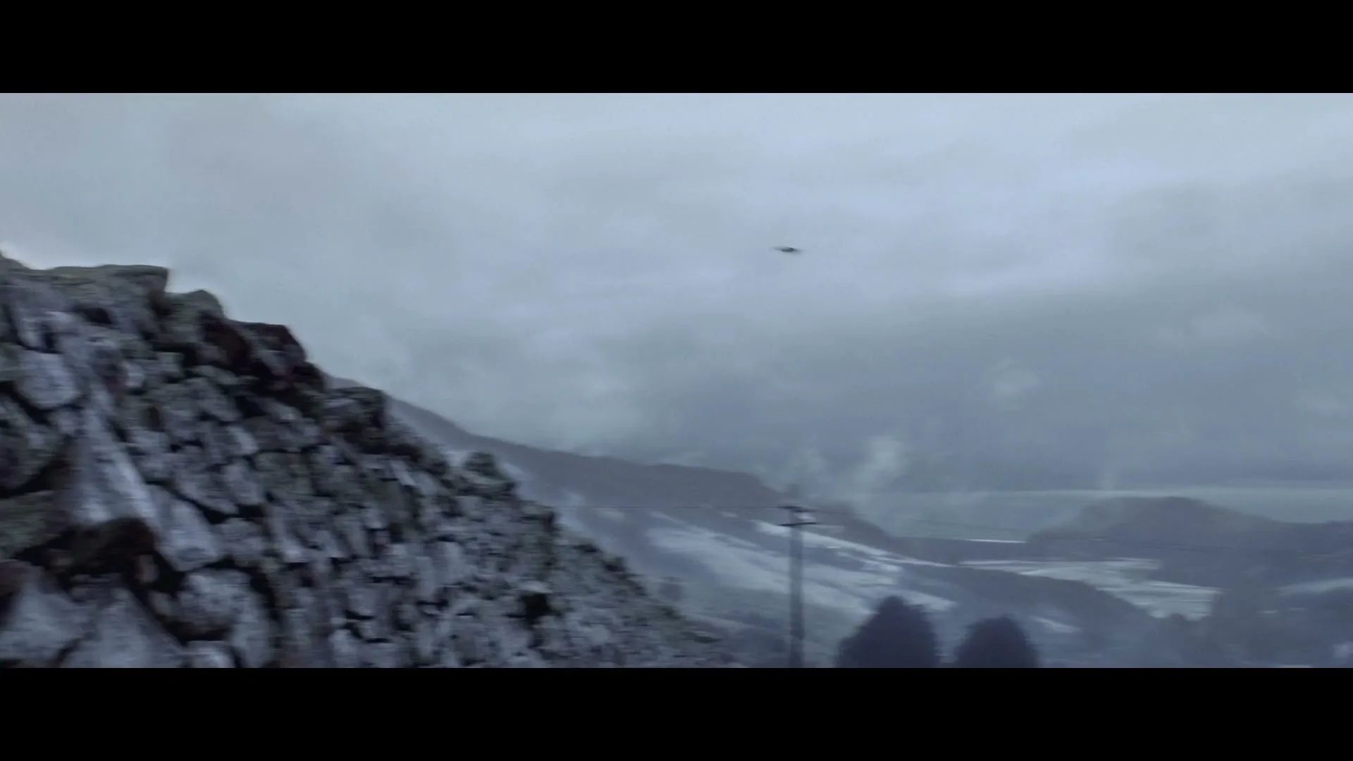 A wide shot of a stone wall on the left, with a vast, misty, snow-dusted mountain landscape and a body of water in the background under a cloudy sky.