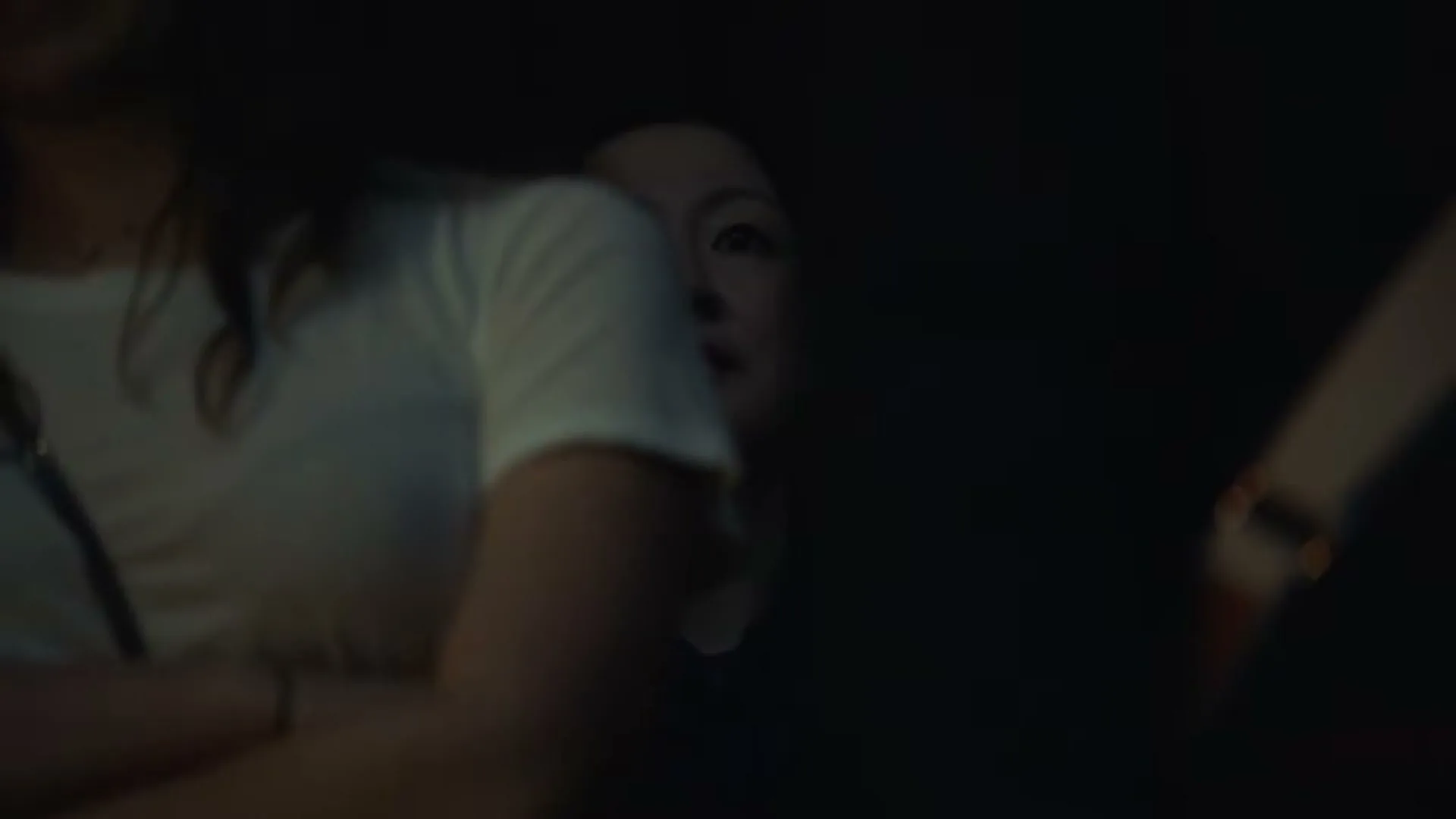 A woman with dark hair and a white shirt is partially visible in the foreground, while another woman with dark hair looks on in the background, in a dimly lit interior.