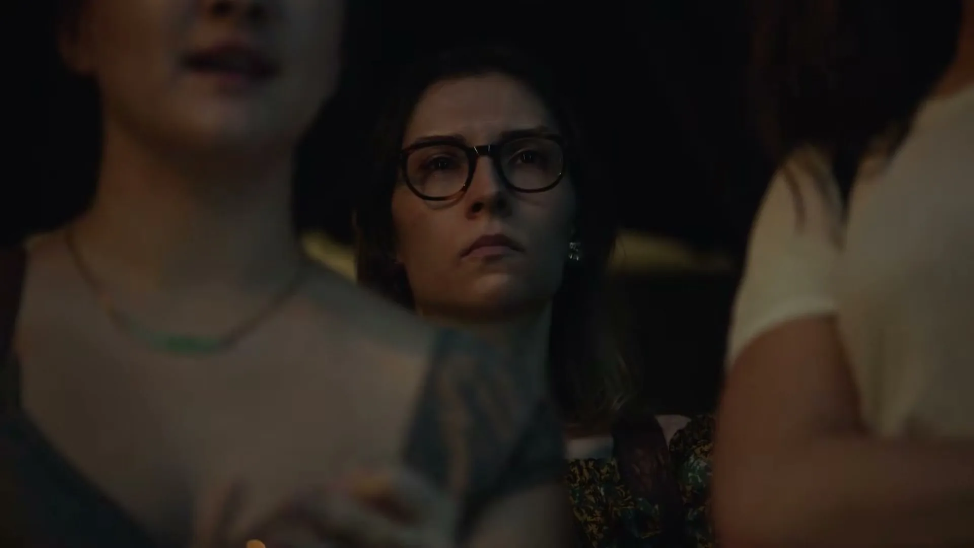 A young woman with glasses looks intently forward, framed by two blurry figures, in a dimly lit interior setting.